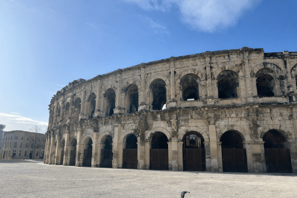 "Vue extérieure des arènes de Nîmes, amphithéâtre romain du Ier siècle, avec ses arches en pierre superposées et sa façade monumentale sous un ciel bleu."